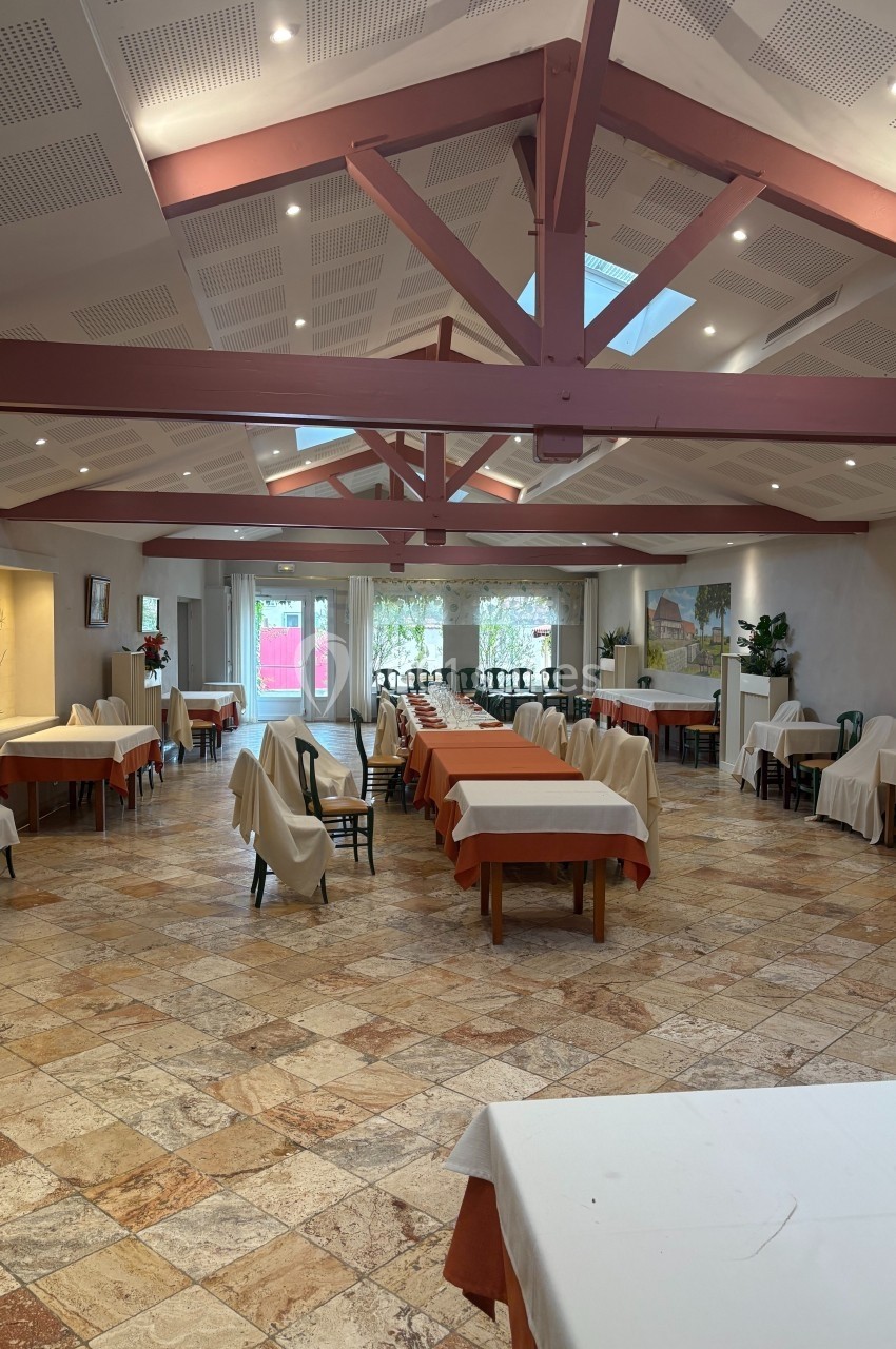 Salle lumineuse avec tables dressées, chaises alignées, sol en carrelage beige et poutres apparentes au plafond.