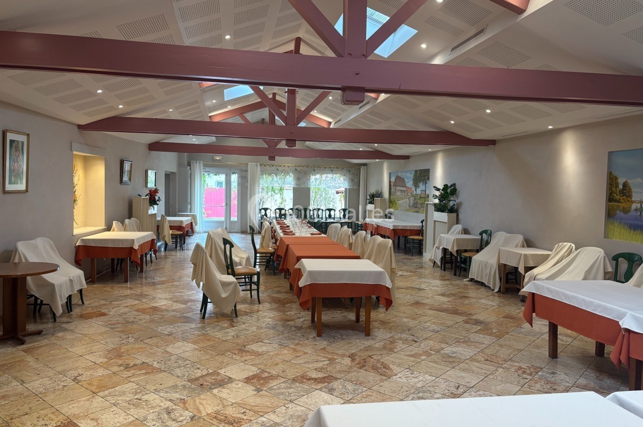 Salle de restaurant lumineuse avec tables dressées, chaises empilées au fond et poutres apparentes au plafond.