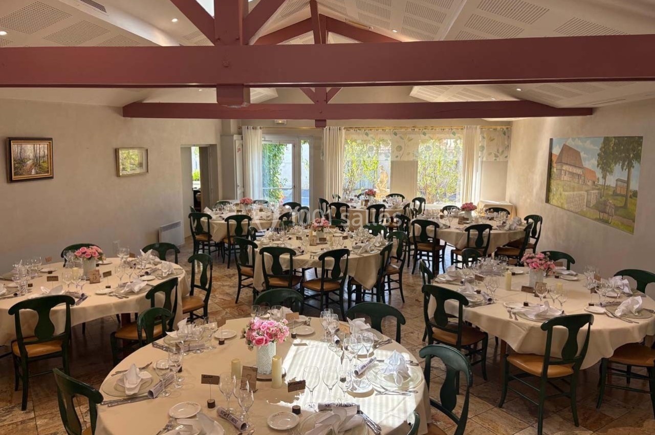 Salle de réception lumineuse avec tables rondes dressées, chaises en bois et décoration florale.
