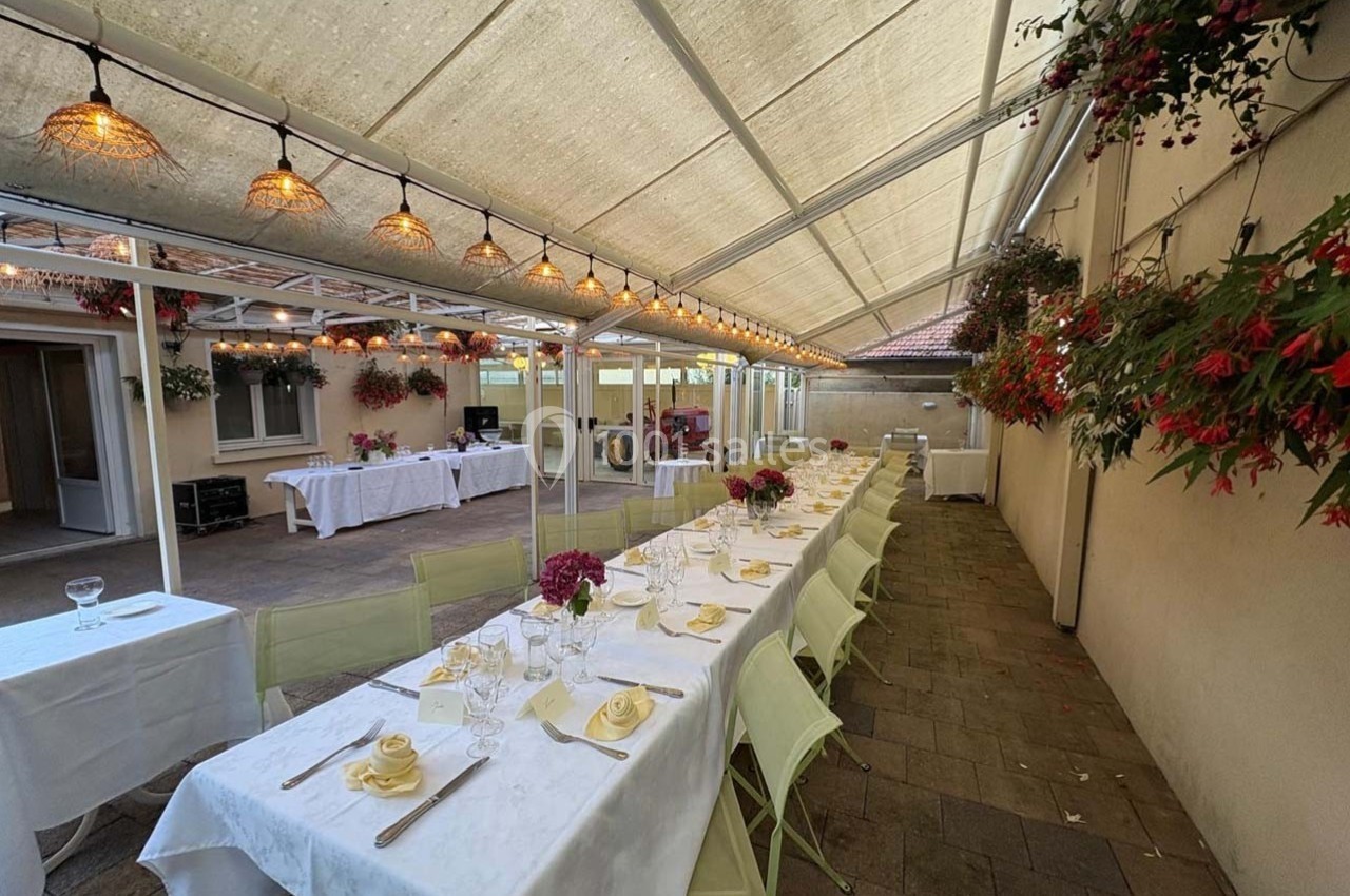 Table dressée sous une véranda lumineuse avec des chaises vertes, décorée de fleurs suspendues et éclairée par des…