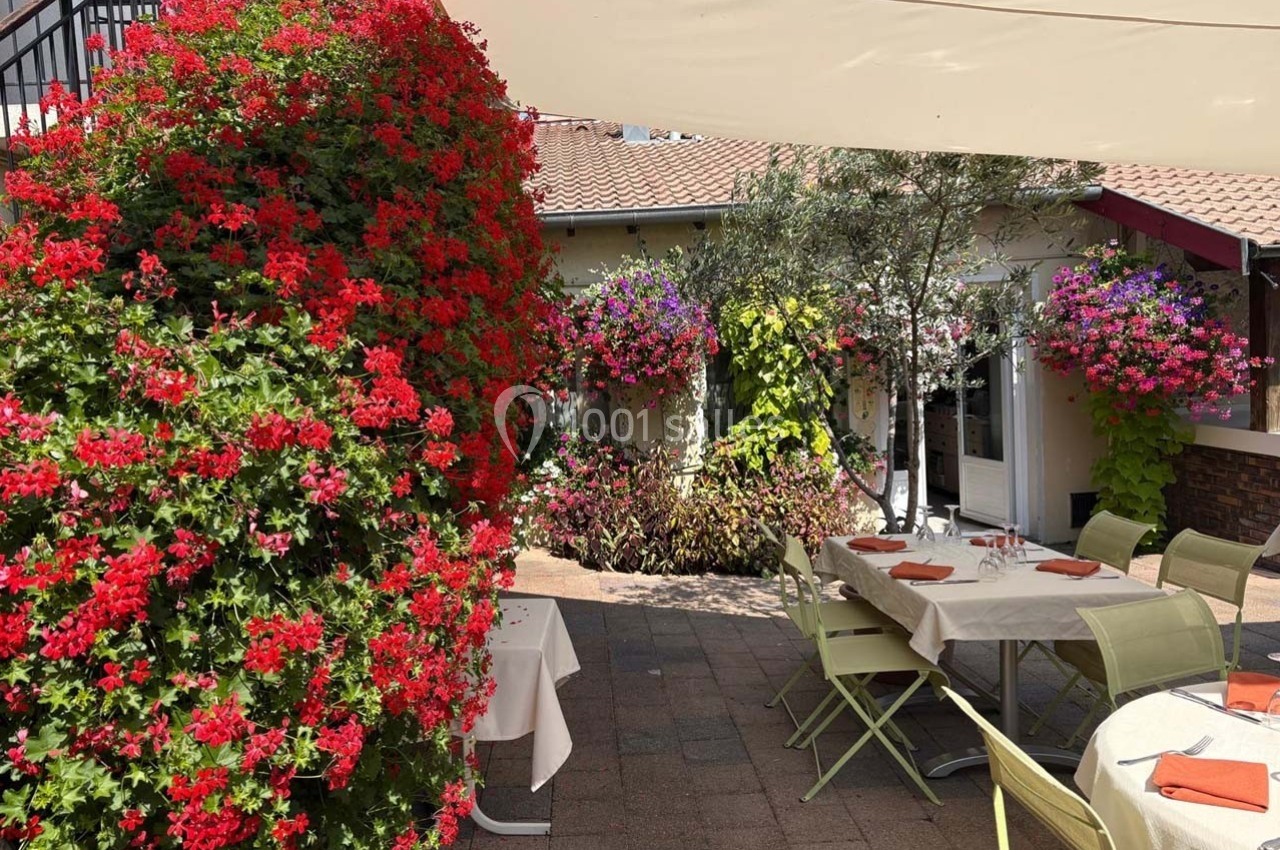 Terrasse extérieure avec tables dressées, entourée de murs fleuris et d'une grande haie de géraniums rouges.