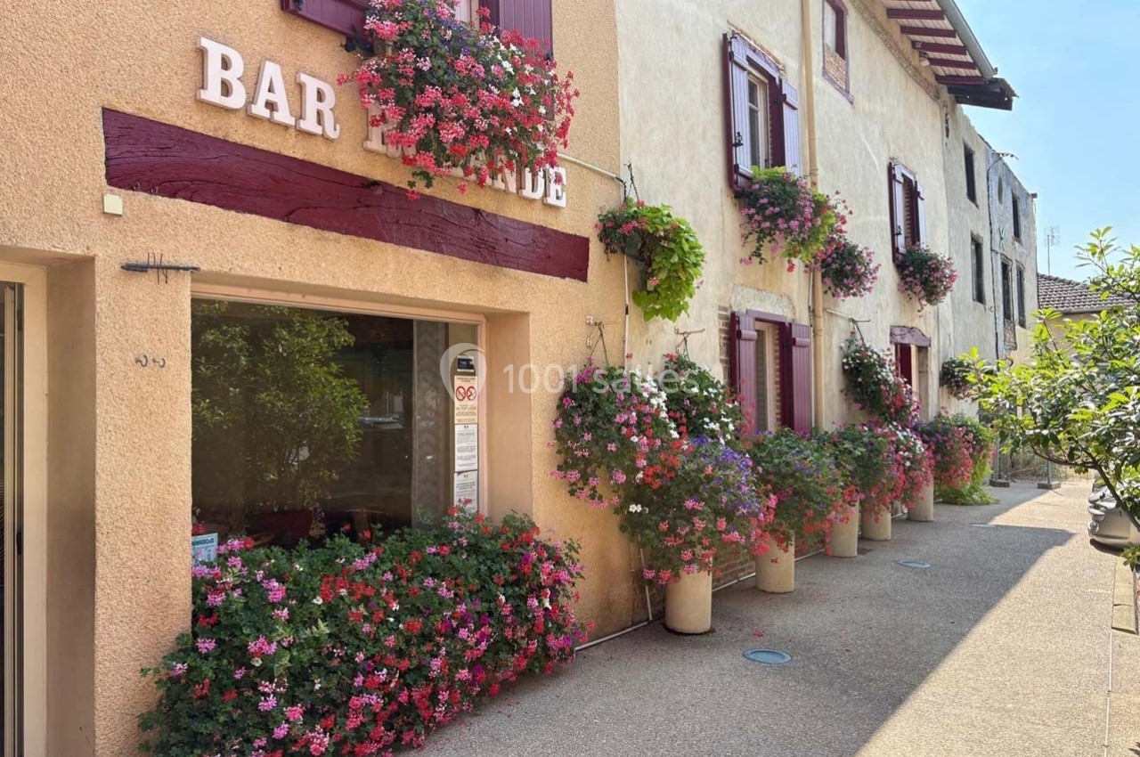 Façade d'un bar et de maisons décorées de volets rouges et de nombreuses fleurs en pots et jardinières.