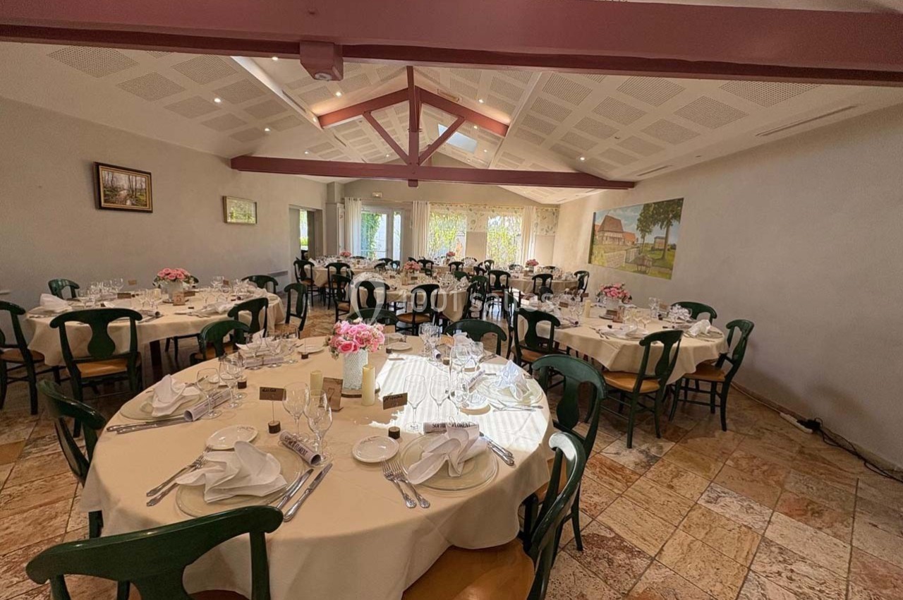 Salle de réception lumineuse avec tables rondes dressées, nappes blanches, chaises en bois et décorations florales.