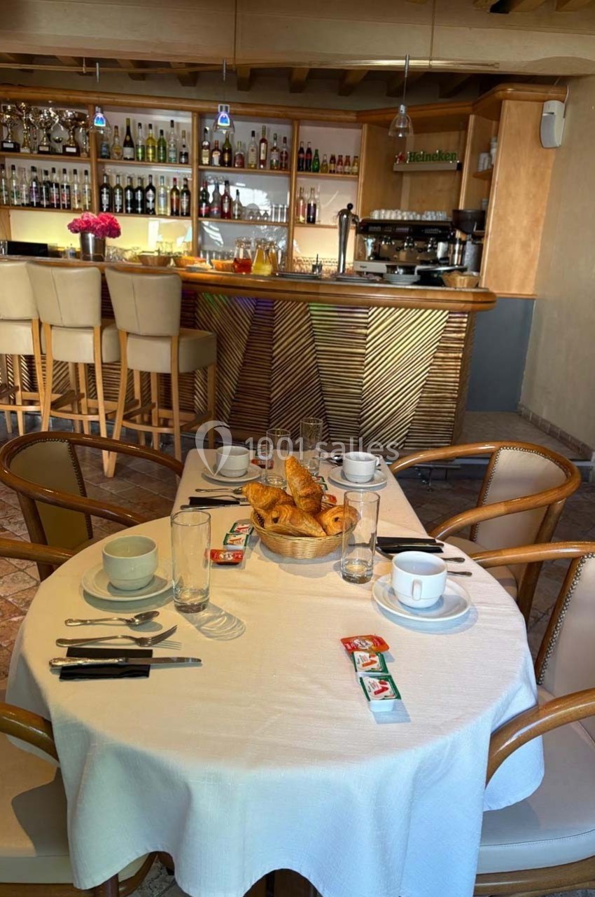 Table dressée pour le petit-déjeuner avec croissants, tasses et couverts, devant un bar bien approvisionné.