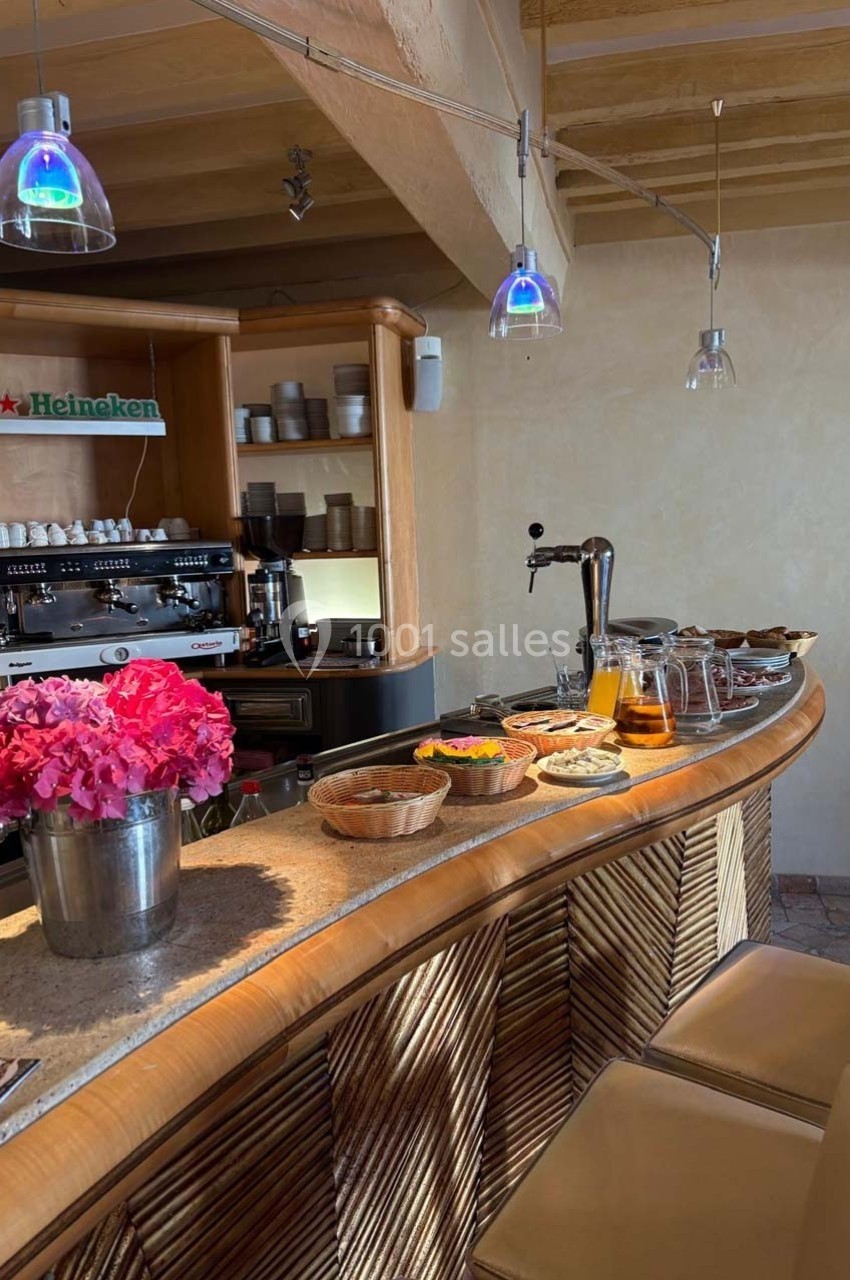 Comptoir de bar en bois avec machine à café, tireuse à bière, viennoiseries, fruits et vase de fleurs roses.