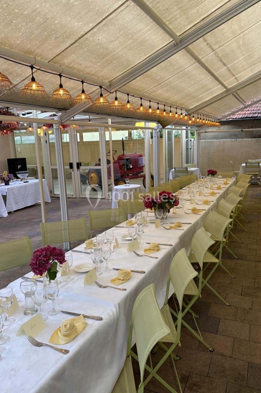 Grande table dressée avec nappes blanches, chaises vertes et décorations florales, sous une pergola lumineuse.