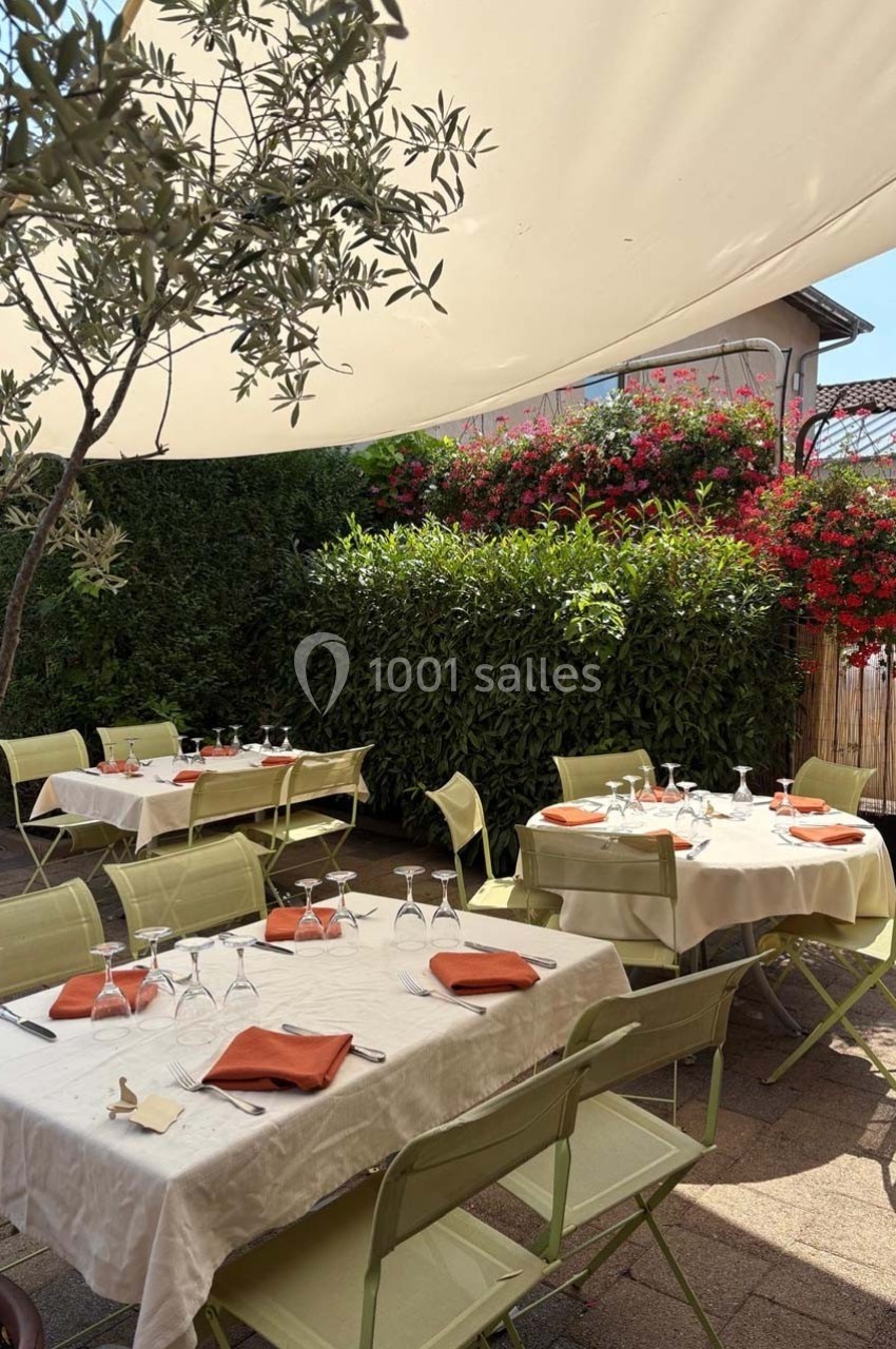 Terrasse extérieure avec tables dressées sous une toile d'ombrage, entourée de verdure et de fleurs colorées.