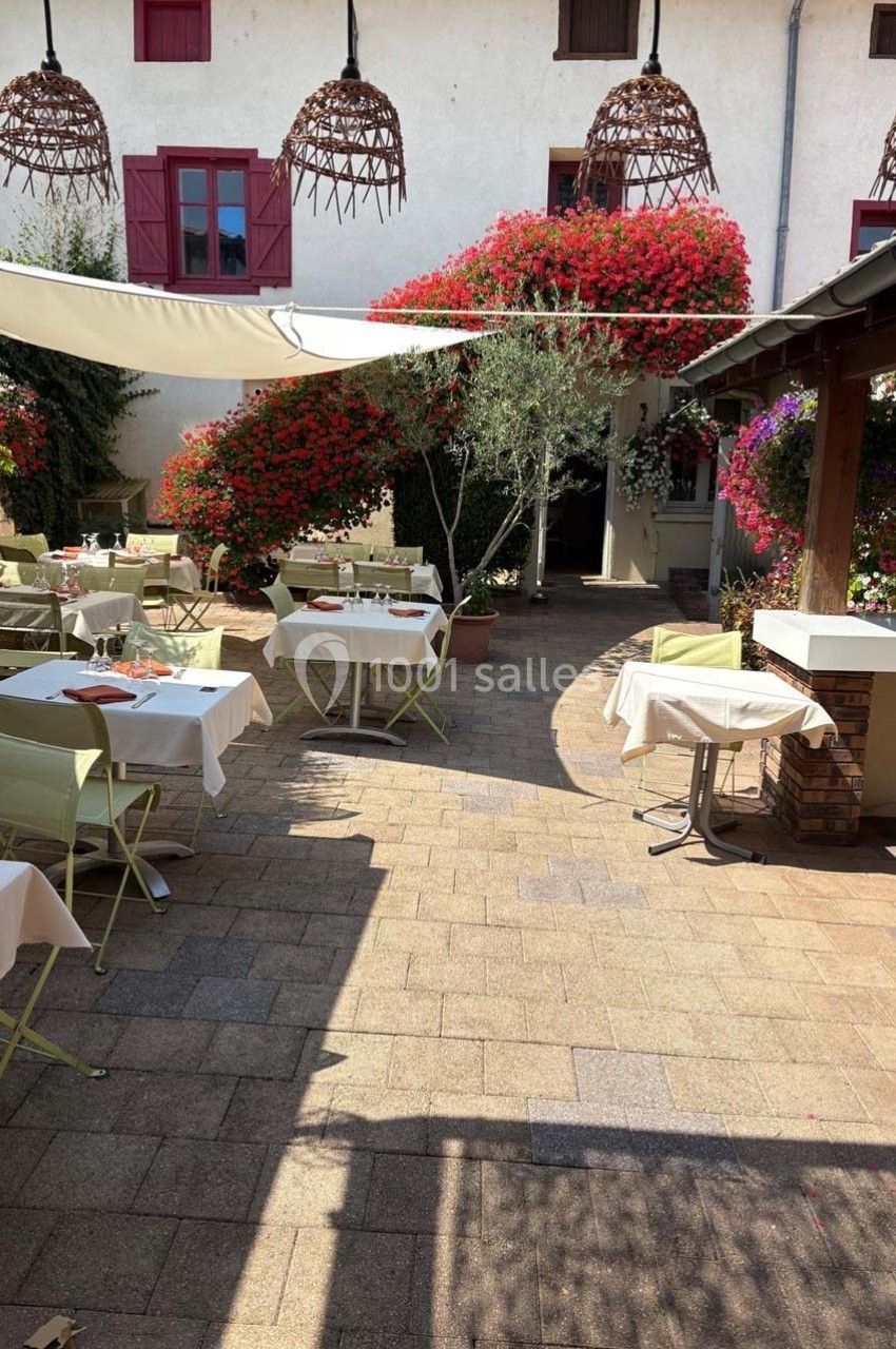 Terrasse d'un restaurant avec tables dressées, chaises vertes, fleurs colorées et pergola dans une cour ensoleillée.