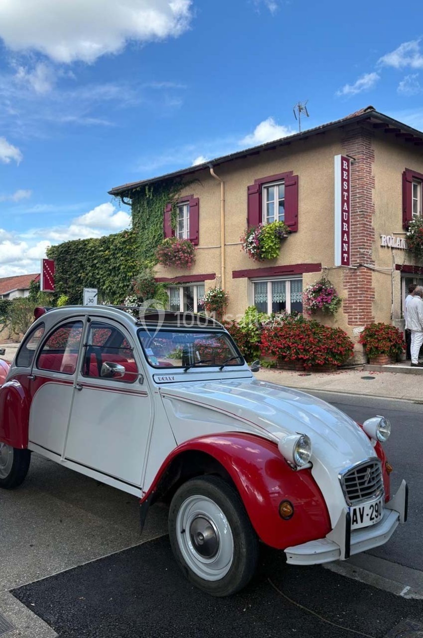 Une voiture Citroën 2CV rouge et blanche est garée devant un restaurant à façade fleurie dans un village.