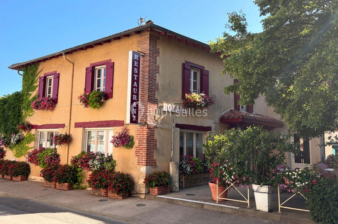 Façade d'un restaurant traditionnel avec murs jaunes, volets rouges et nombreuses fleurs en pots et jardinières.