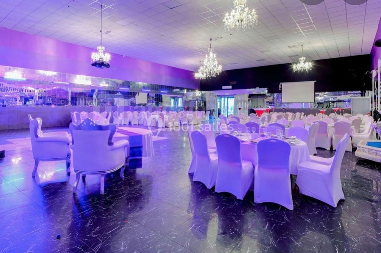 Salle de réception élégante avec tables rondes dressées, chaises blanches et lustres suspendus.
