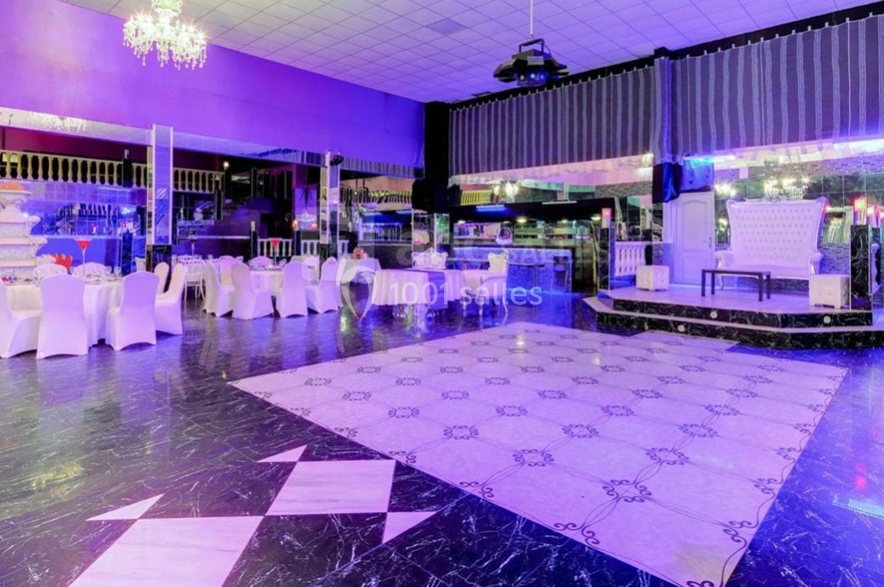 Salle de réception spacieuse avec tables dressées, chaises blanches, piste de danse et éclairage violet.