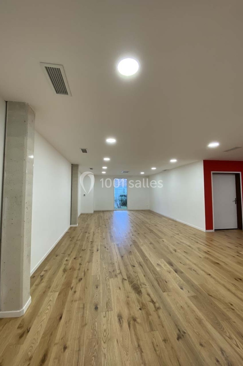Salle vide avec parquet en bois clair, murs blancs, un mur rouge, éclairage encastré et une porte vitrée au fond.