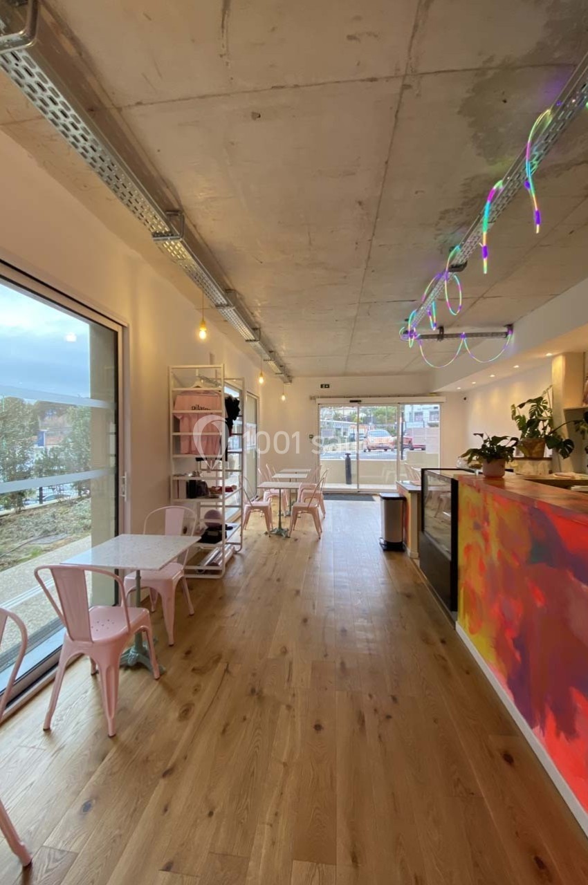 Intérieur lumineux d'un café moderne avec des tables, chaises roses, comptoir coloré et vue sur l'extérieur.