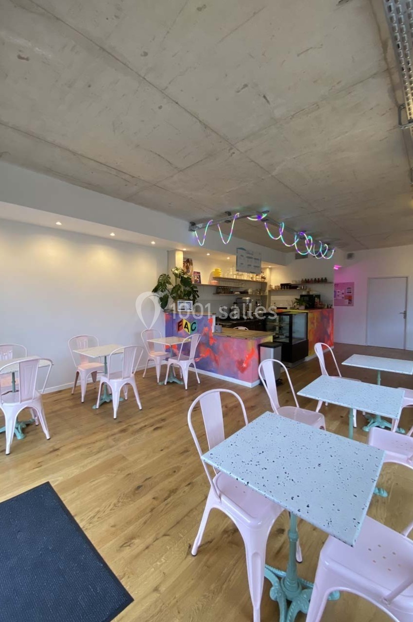 Salle de café moderne avec tables en terrazzo, chaises roses, comptoir coloré et éclairage néon sous un plafond brut.