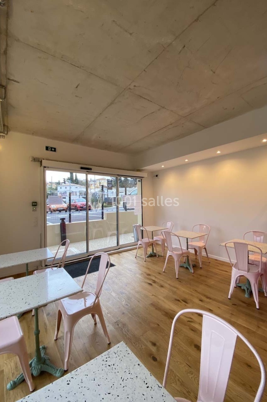 Salle lumineuse avec tables en terrazzo, chaises roses et grandes baies vitrées donnant sur l'extérieur.