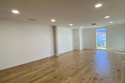 Salle vide avec parquet en bois clair, murs blancs, éclairage encastré au plafond et grande baie vitrée donnant sur l…