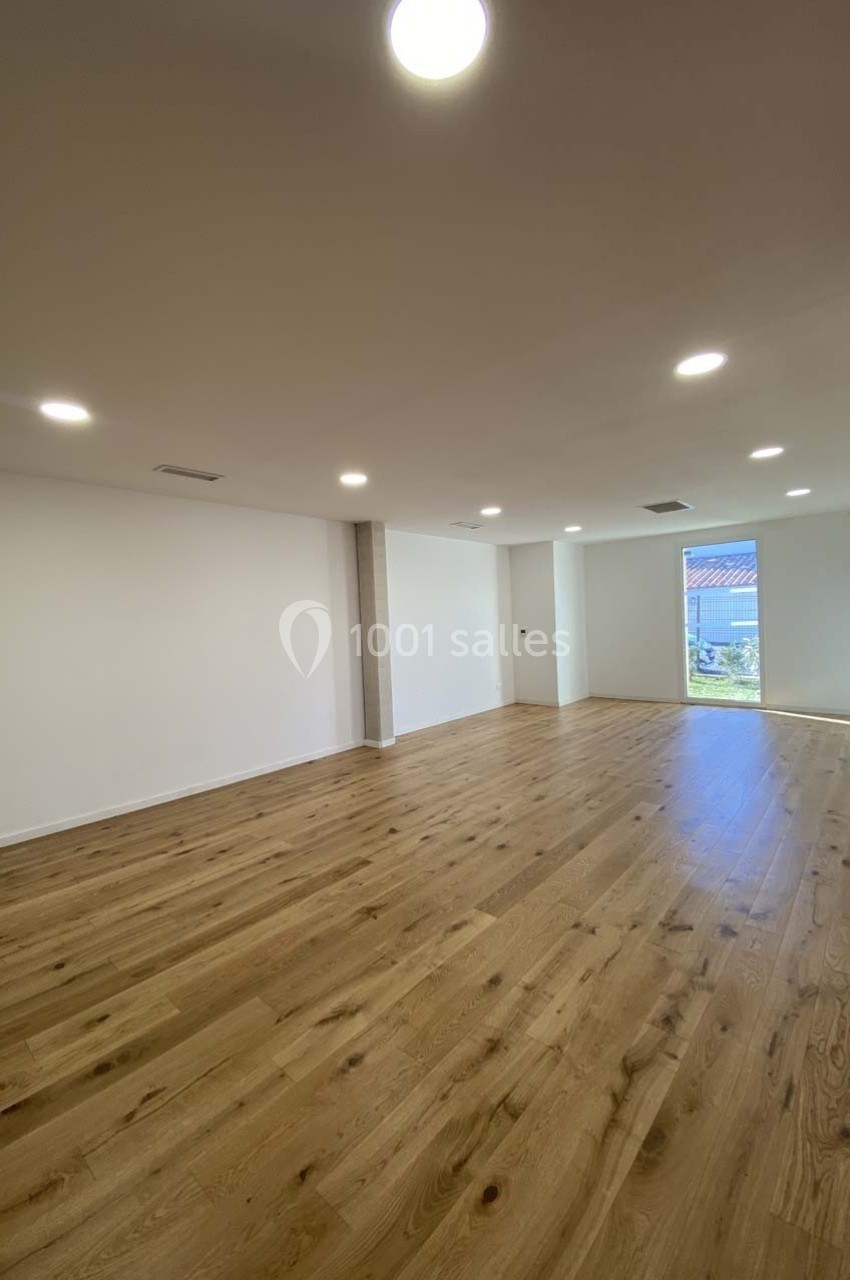 Salle vide avec parquet en bois clair, murs blancs, éclairage encastré au plafond et grande fenêtre donnant sur l'extérieur.