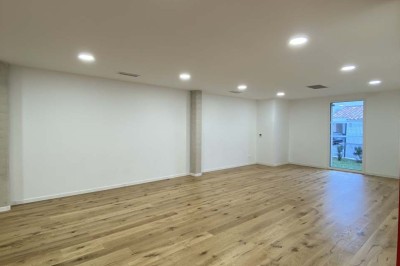 Salle vide avec parquet en bois clair, murs blancs, éclairage encastré au plafond et grande baie vitrée donnant sur l…