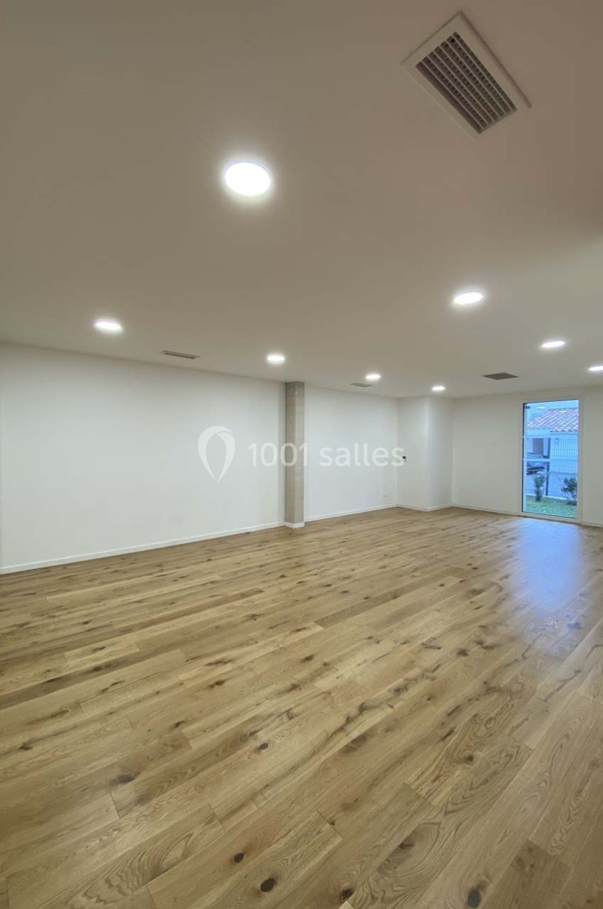 Salle vide avec parquet en bois clair, murs blancs, éclairage encastré au plafond et grande baie vitrée donnant sur l…