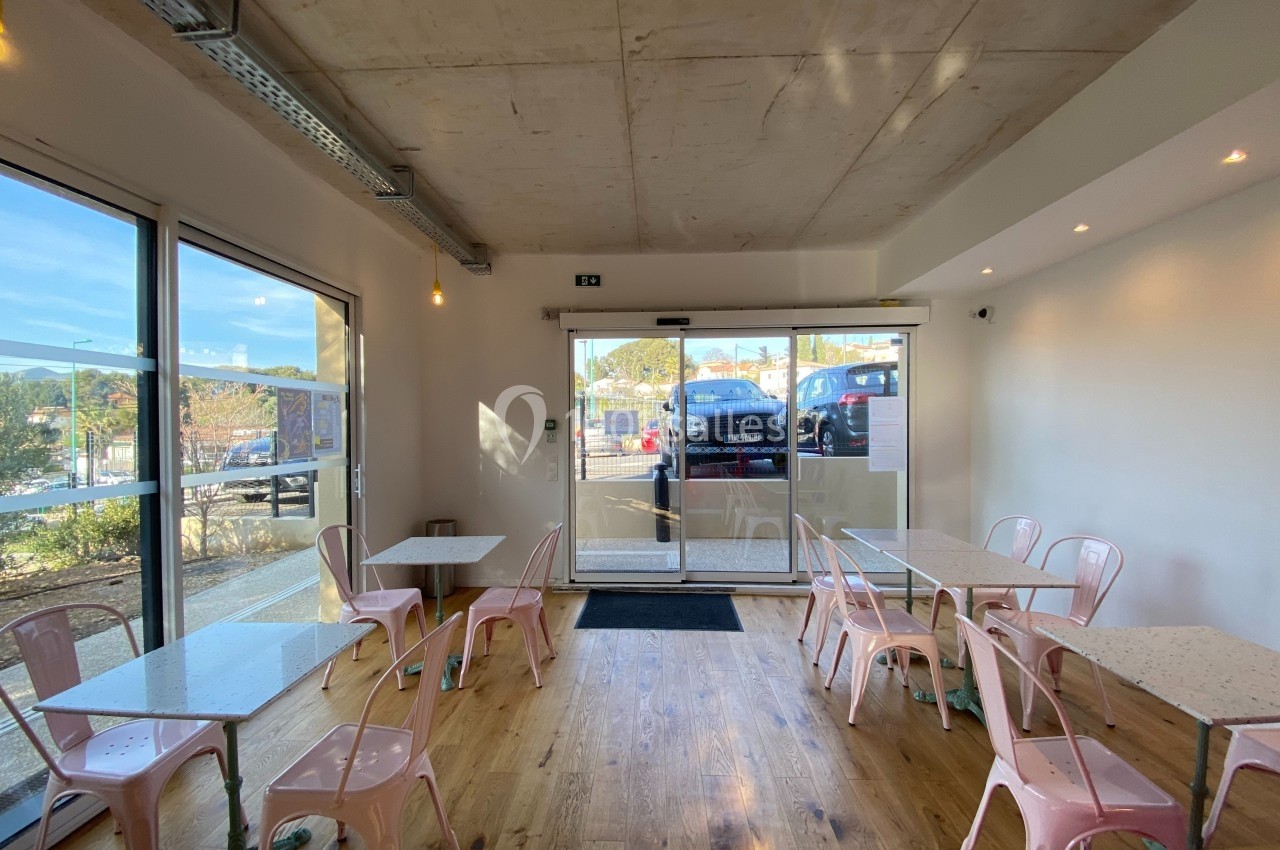 Salle de café lumineuse avec tables et chaises roses, grandes baies vitrées donnant sur un parking.