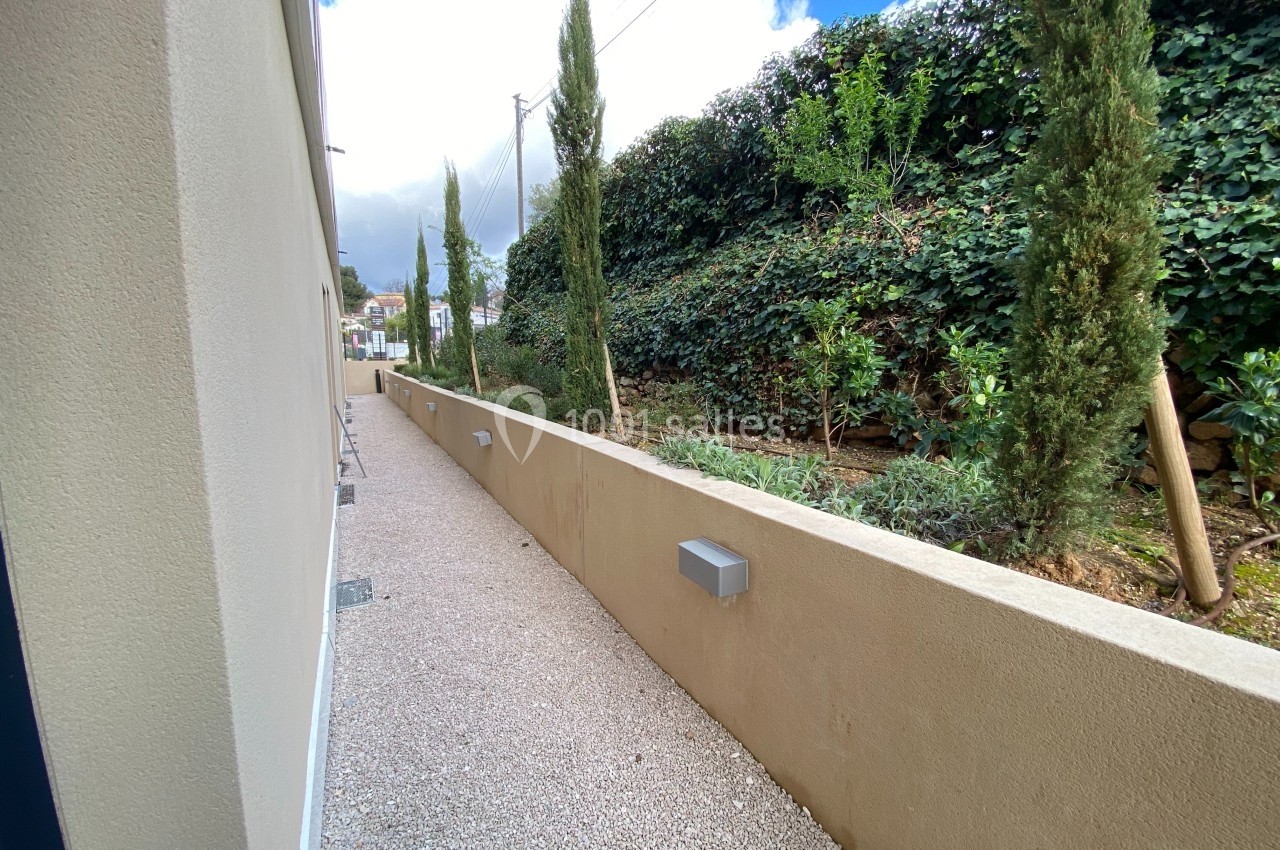 Allée extérieure en gravier longeant un mur beige, bordée de cyprès et d'une haie végétale sur terrain en pente.