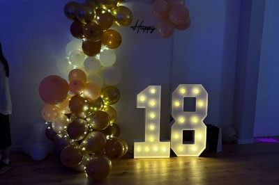 Décoration lumineuse avec le chiffre 18 éclairé et des ballons dorés et blancs dans une pièce sombre.