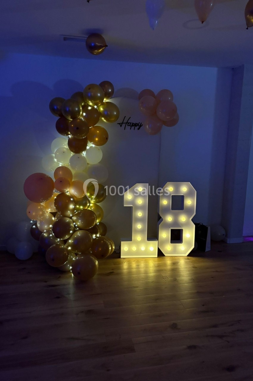 Décoration lumineuse avec le chiffre 18 éclairé et des ballons dorés et blancs dans une pièce sombre.