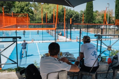 Miniature Location salle Simiane-Collongue (Bouches-du-Rhône) - Restaurant du Cercle #8 Terrain de padel extérieur avec sol bleu, clôtures en verre et grillage, entouré d'arbres et de filets orange.
