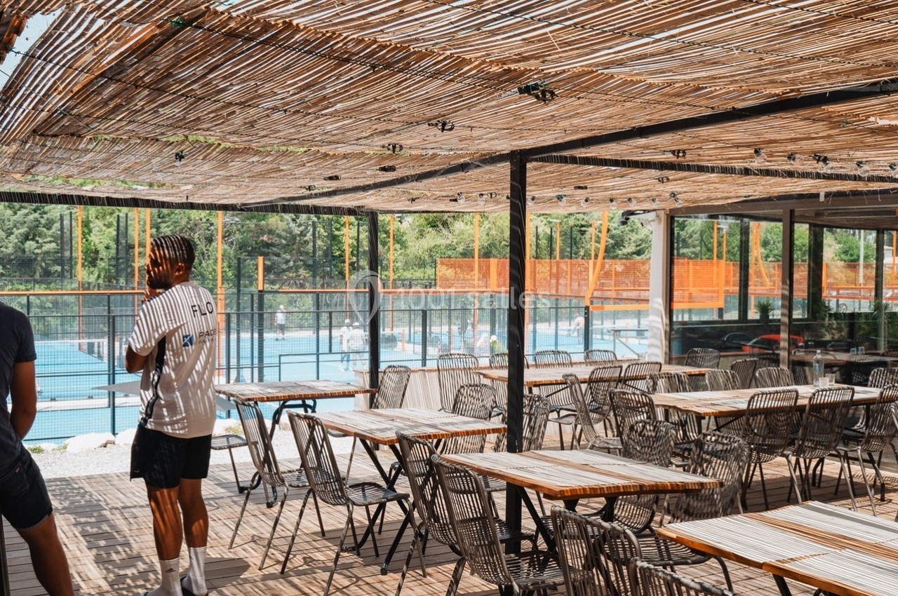 Terrasse ombragée avec des tables et des chaises, donnant sur une piscine entourée de végétation et de clôtures en bois.
