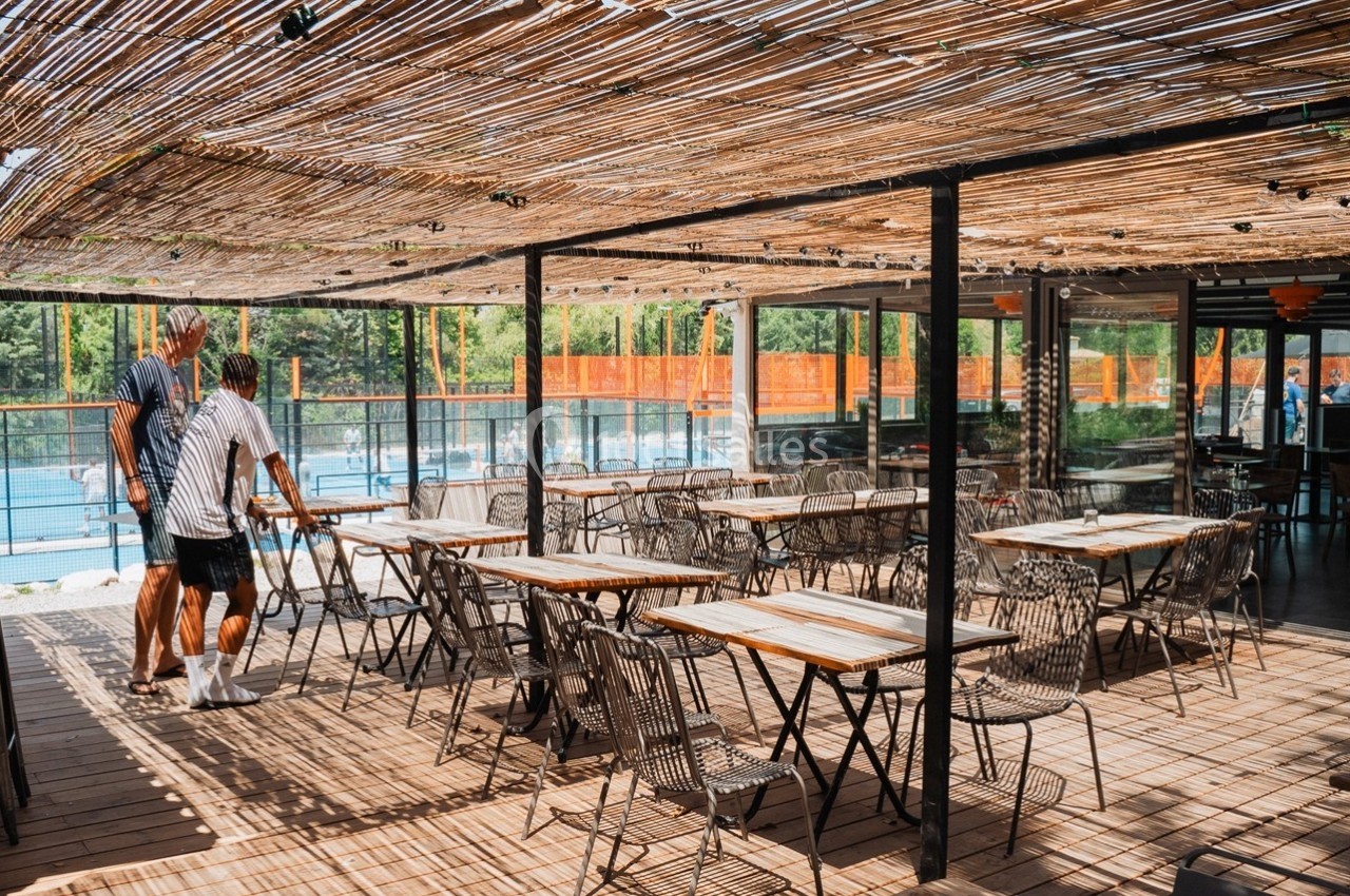 Terrasse en bois avec tables et chaises sous une pergola en bambou, donnant sur un terrain de sport extérieur.