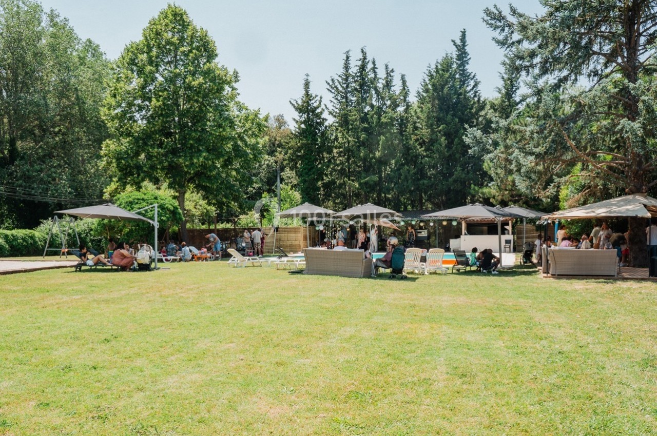Jardin avec pelouse, parasols, chaises et personnes rassemblées près de structures en bois sous un ciel dégagé.