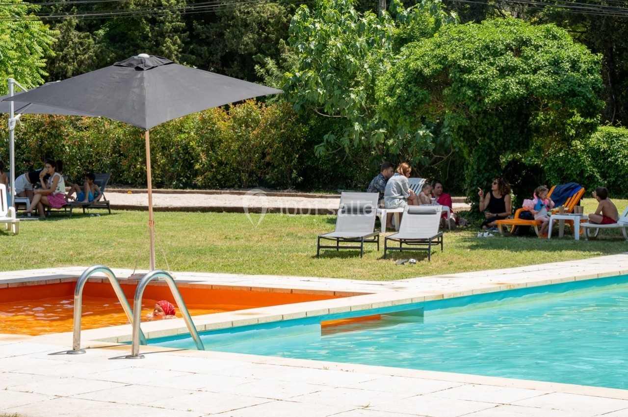 Piscine extérieure avec échelle, parasol noir et personnes assises sur des chaises longues dans un espace vert.