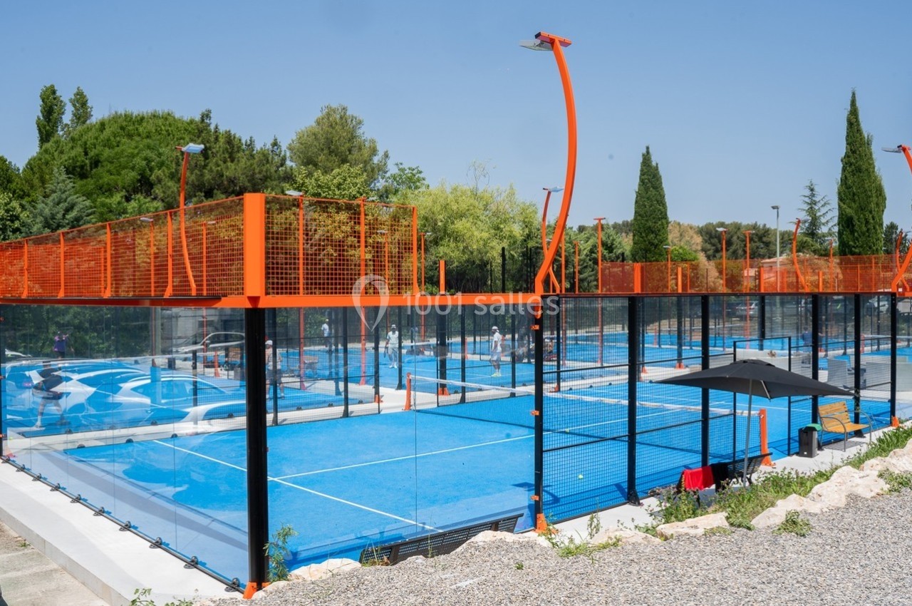 Terrains de padel extérieurs avec sol bleu, entourés de grillages orange, situés dans un environnement arboré.