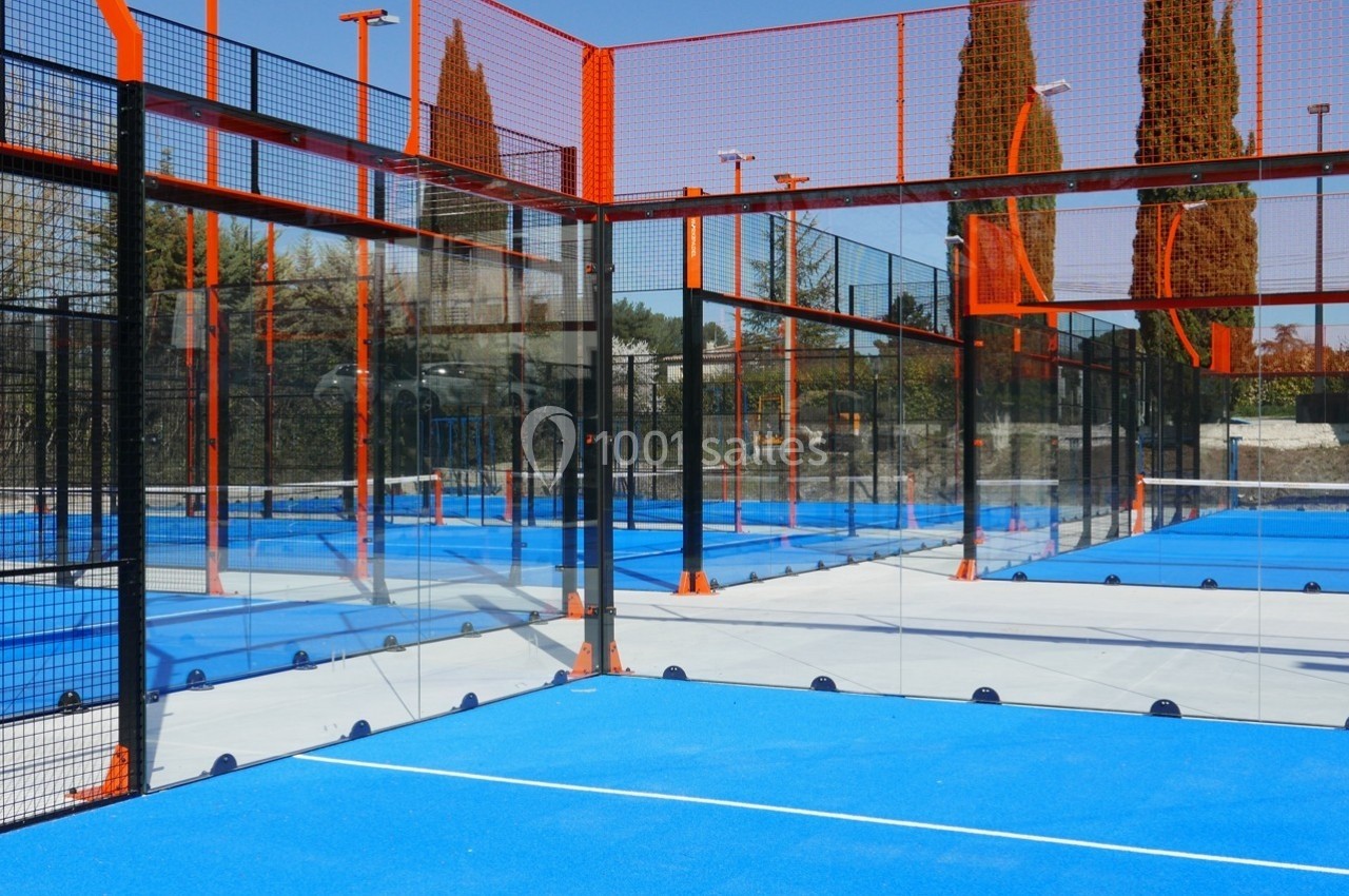 Terrain de padel extérieur avec sol bleu, clôtures en verre et grillage, entouré d'arbres et de filets orange.