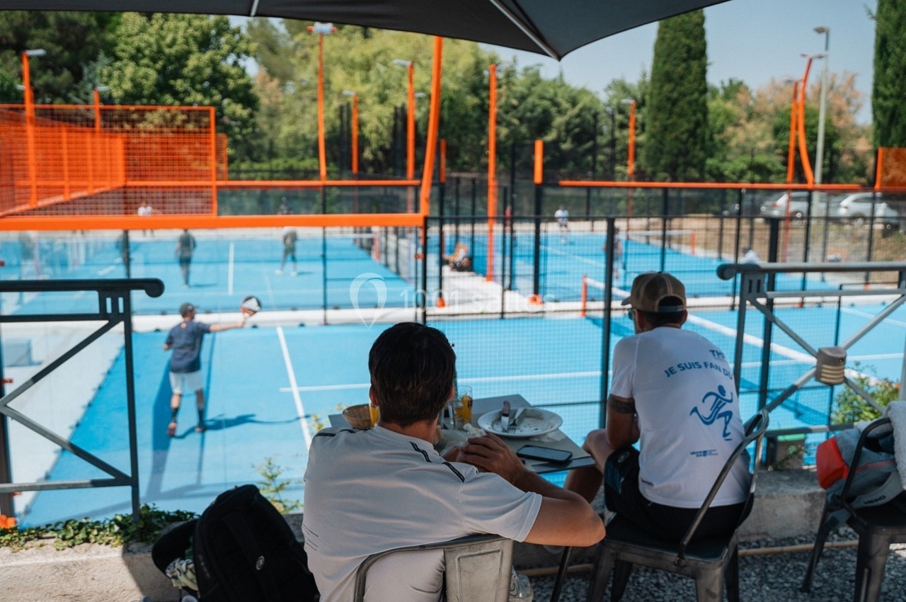 Deux hommes assis à une table en terrasse regardent des joueurs sur des terrains de padel en plein air.