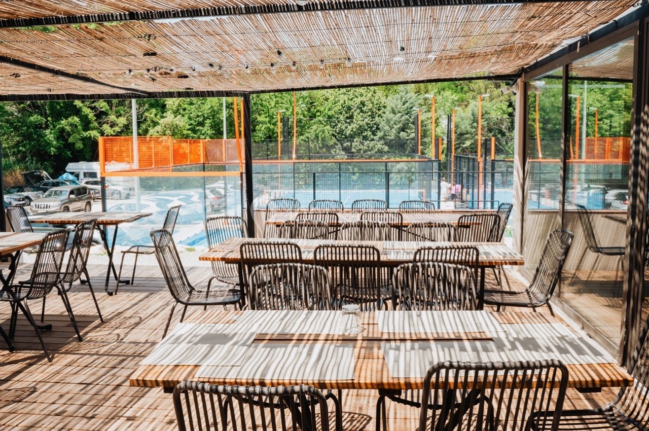 Terrasse en bois ombragée par une pergola en bambou, avec tables et chaises, vue sur une piscine et des arbres.