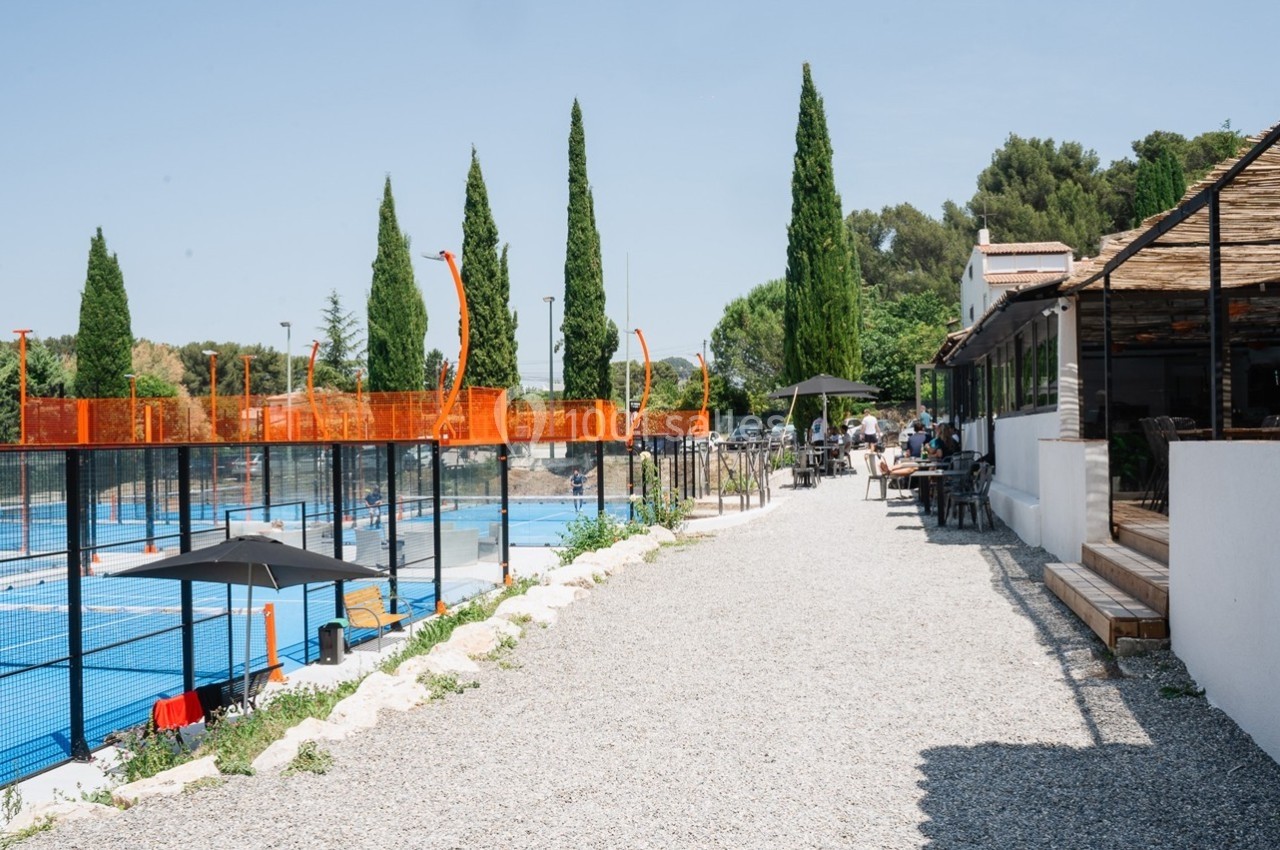 Terrasse ombragée près d'un terrain de padel entouré de cyprès sous un ciel dégagé.