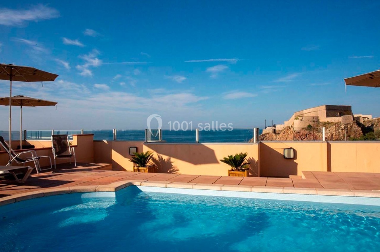 Piscine extérieure avec vue sur la mer et un fort historique en arrière-plan, sous un ciel dégagé.