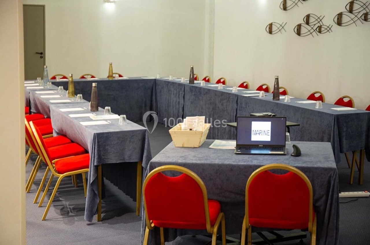 Salle de réunion avec tables en U couvertes de nappes bleues, chaises rouges et un ordinateur portable allumé.