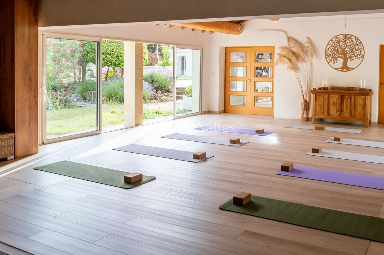 Salle lumineuse avec des tapis de yoga alignés au sol, blocs en bois, grandes baies vitrées donnant sur un jardin.