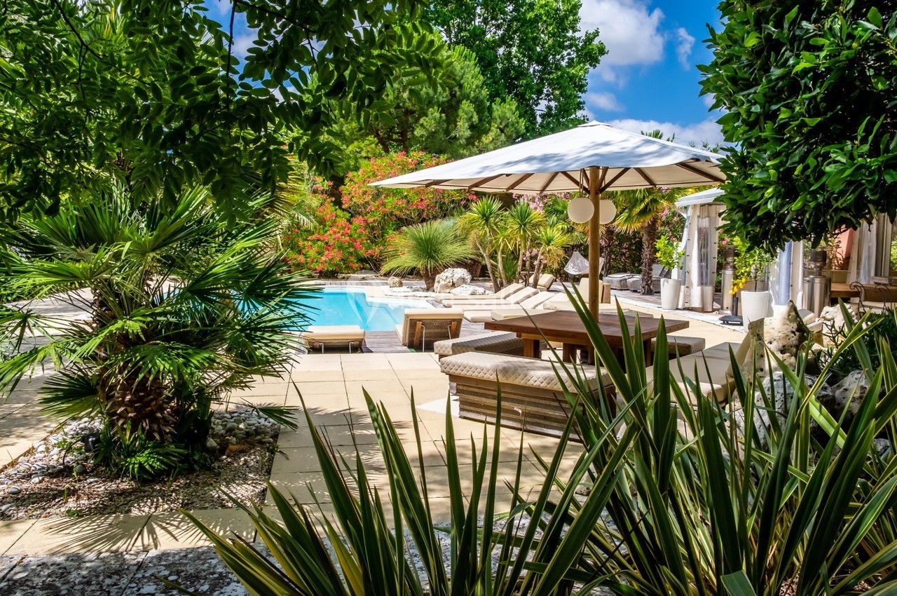 Jardin avec piscine entourée de végétation luxuriante, chaises longues et parasol blanc sur une terrasse ensoleillée.