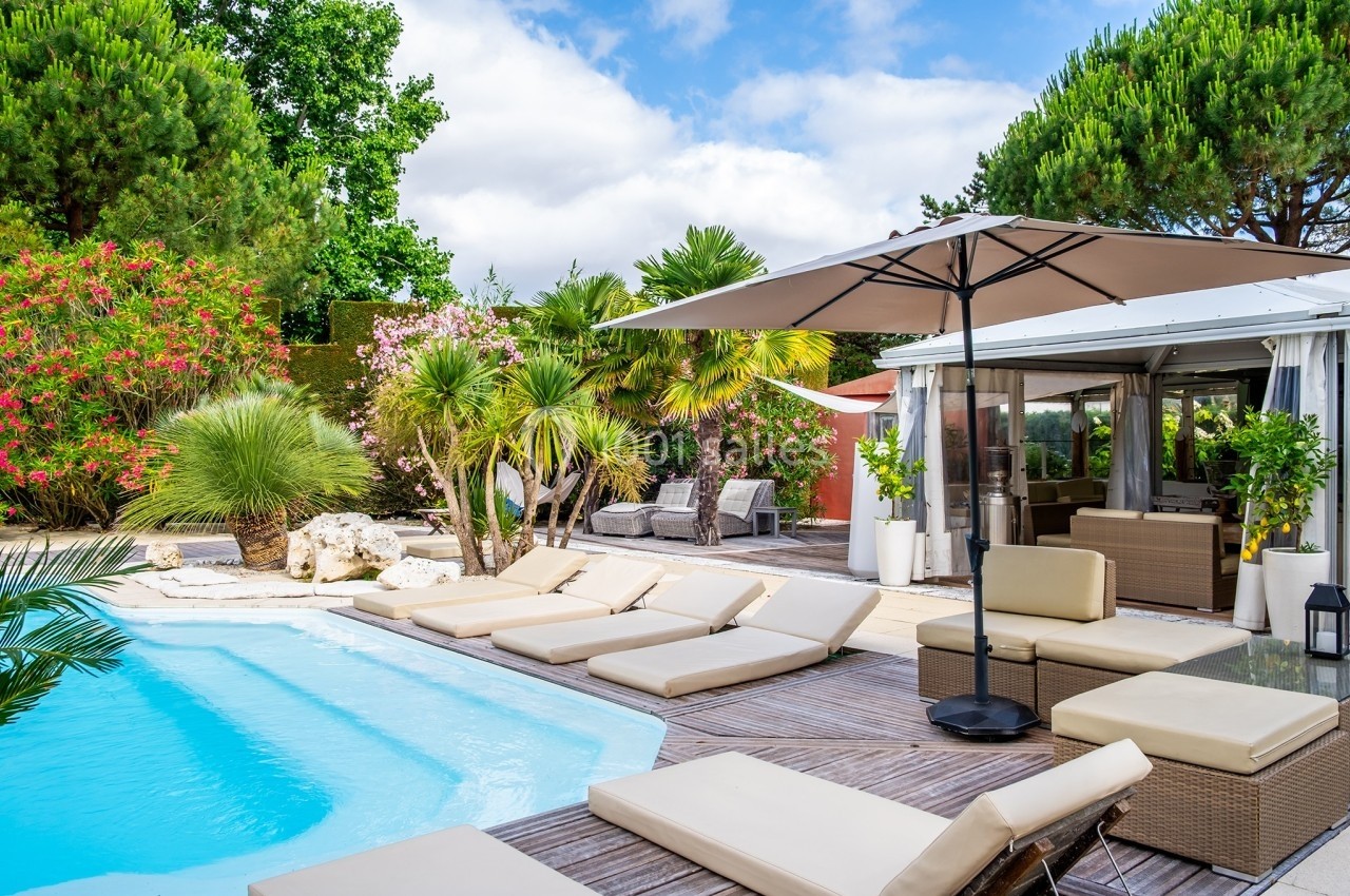 Terrasse avec piscine, transats beiges, parasol et végétation luxuriante sous un ciel partiellement nuageux.