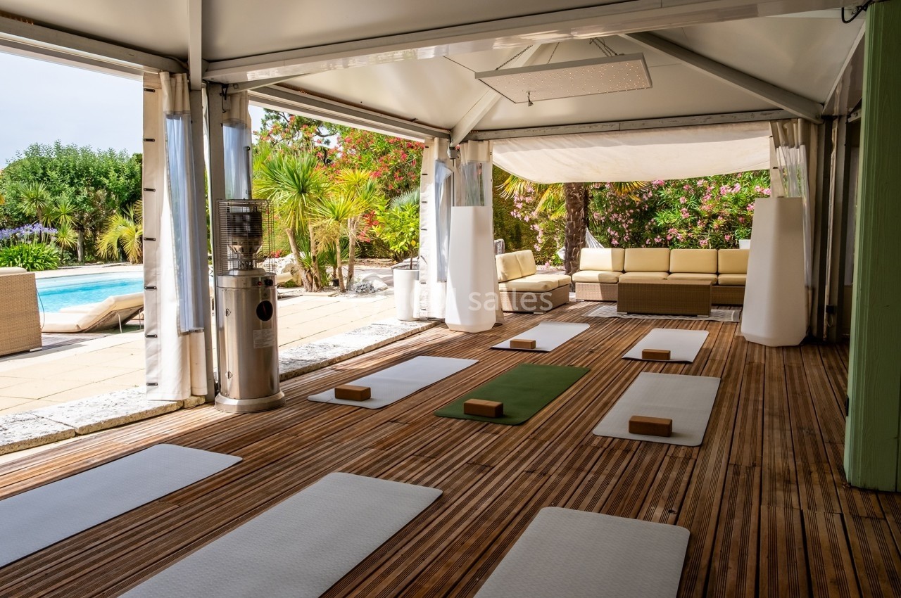 Salle de yoga en plein air avec tapis et briques disposés sur un sol en bois, entourée de végétation et proche d'une piscine.