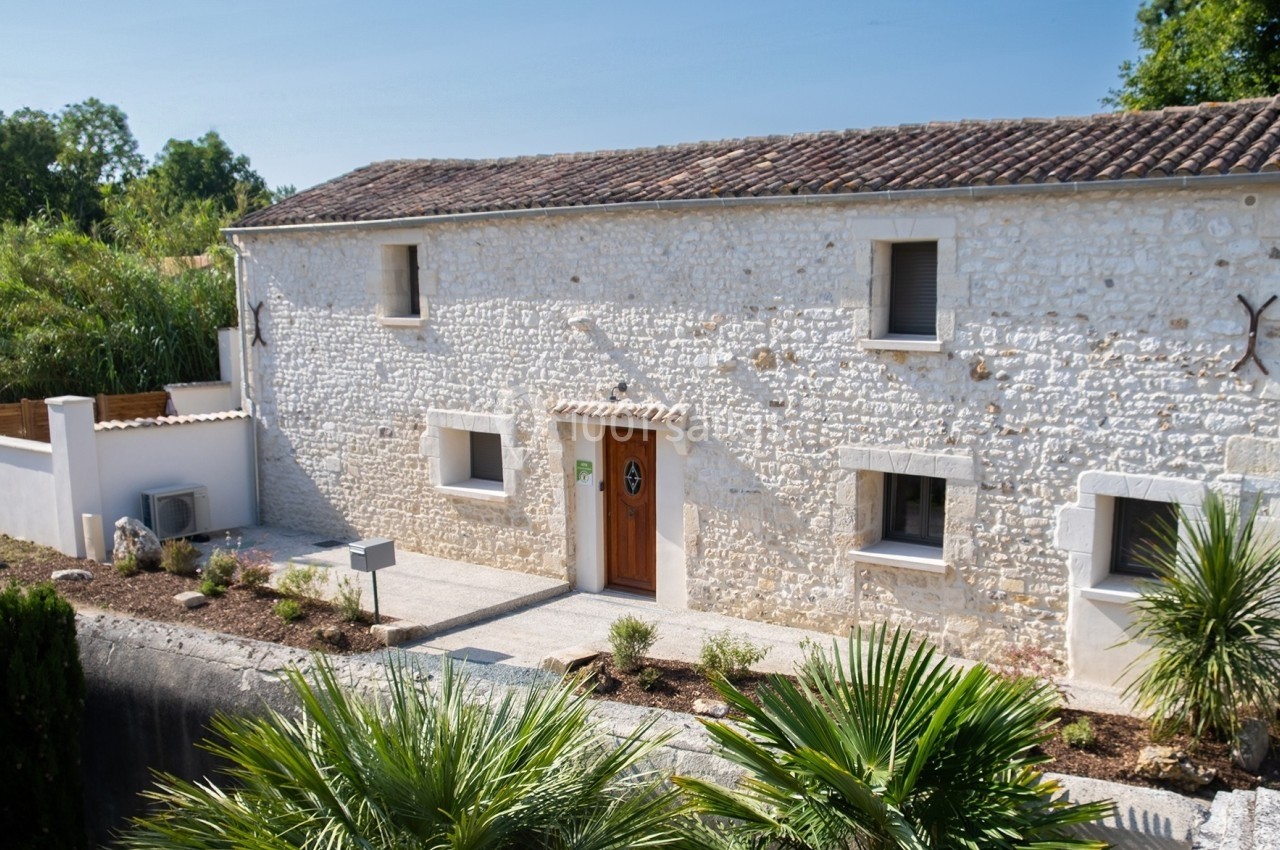 Façade d'une maison en pierre blanche avec toit en tuiles, entourée de végétation et d'un jardin aménagé.