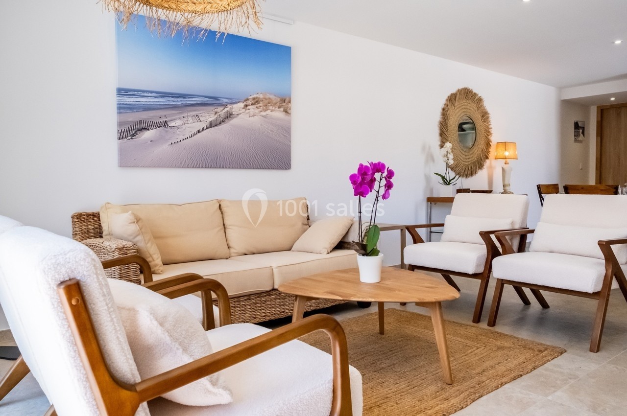 Salon lumineux avec mobilier en bois et rotin, orchidée violette sur une table basse, tableau de dunes en arrière-plan.