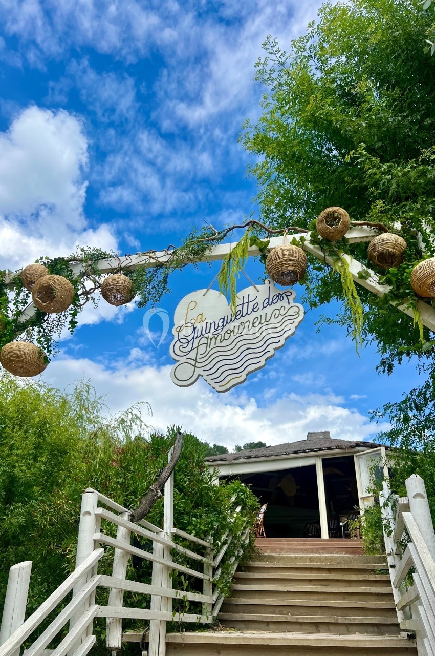 Entrée décorée avec des lanternes et un panneau ’Guinguette des Amoureux’, entourée de verdure sous un ciel bleu.