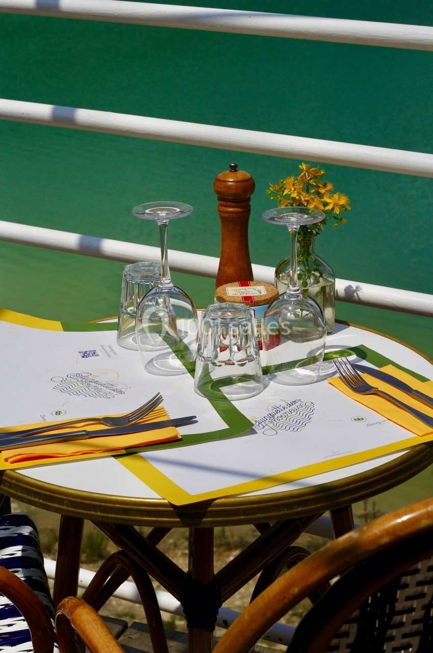 Table dressée en extérieur avec nappes colorées, verres, couverts, poivrier et vase de fleurs, près d'une balustrade blanche.