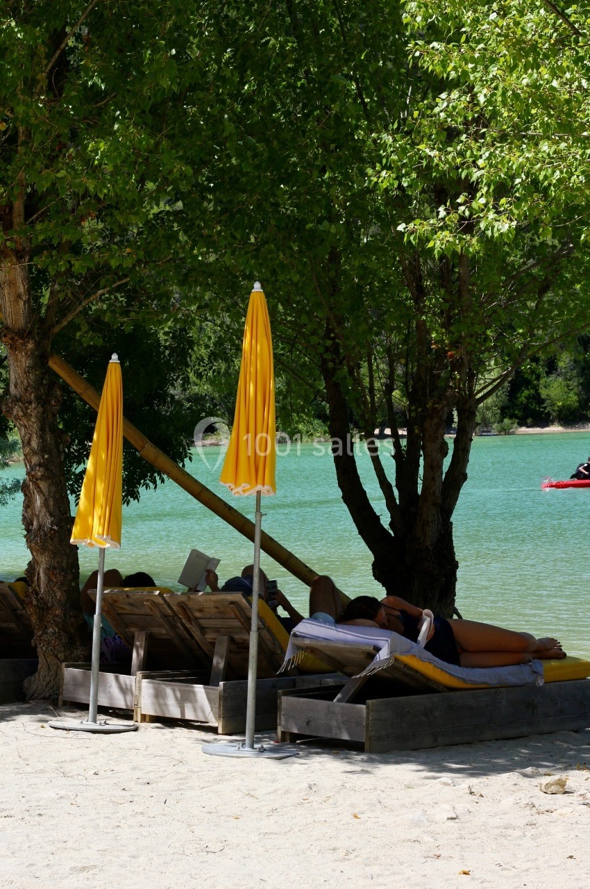 Des transats sous des arbres avec des parasols jaunes, au bord d'un lac aux eaux turquoise.