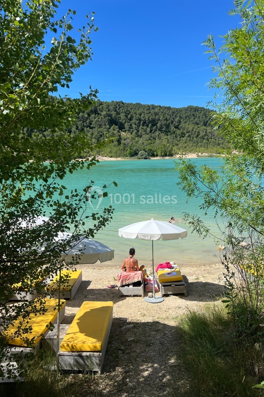 Chaises longues jaunes et parasols blancs au bord d'un lac turquoise entouré de végétation et de collines boisées.